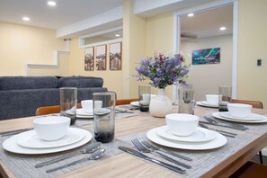 Un comedor con una mesa de madera dispuesta para cuatro personas, platos blancos, cubiertos y vasos transparentes. Un jarrón con flores moradas está sobre la mesa.