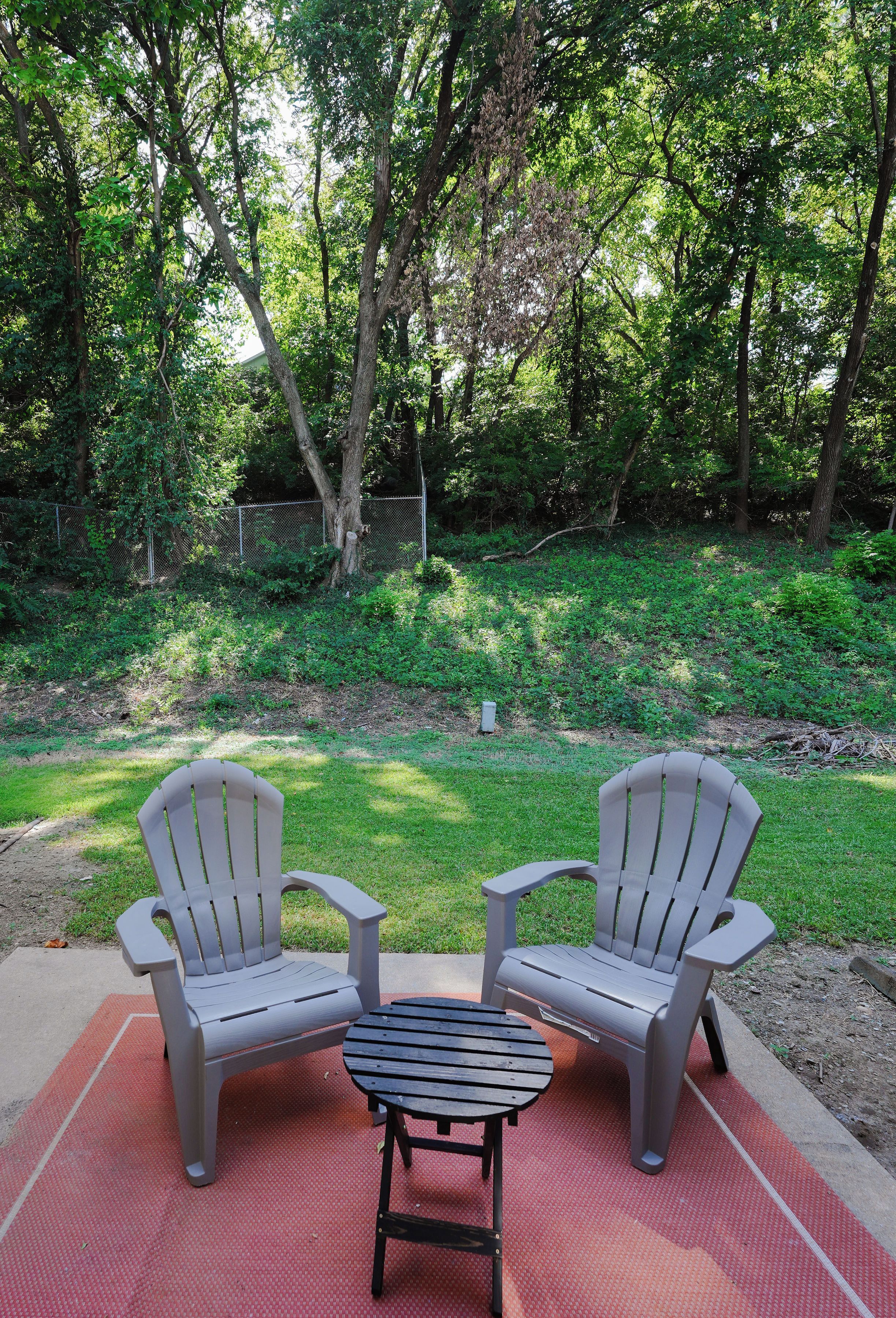 Two Adirondack chairs on a red mat with a small table, surrounded by a grassy area and trees.