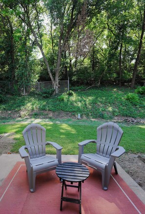 Two Adirondack chairs on a red mat with a small table, surrounded by a grassy area and trees.