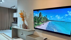 A modern hotel room with a large flat-screen TV displaying a beach scene, a bedside table with a vase of flowers, a bed with a white comforter, and a grey cabinet. - Seaside Studio on the ecological route of Milagres (Porto de Pedras)