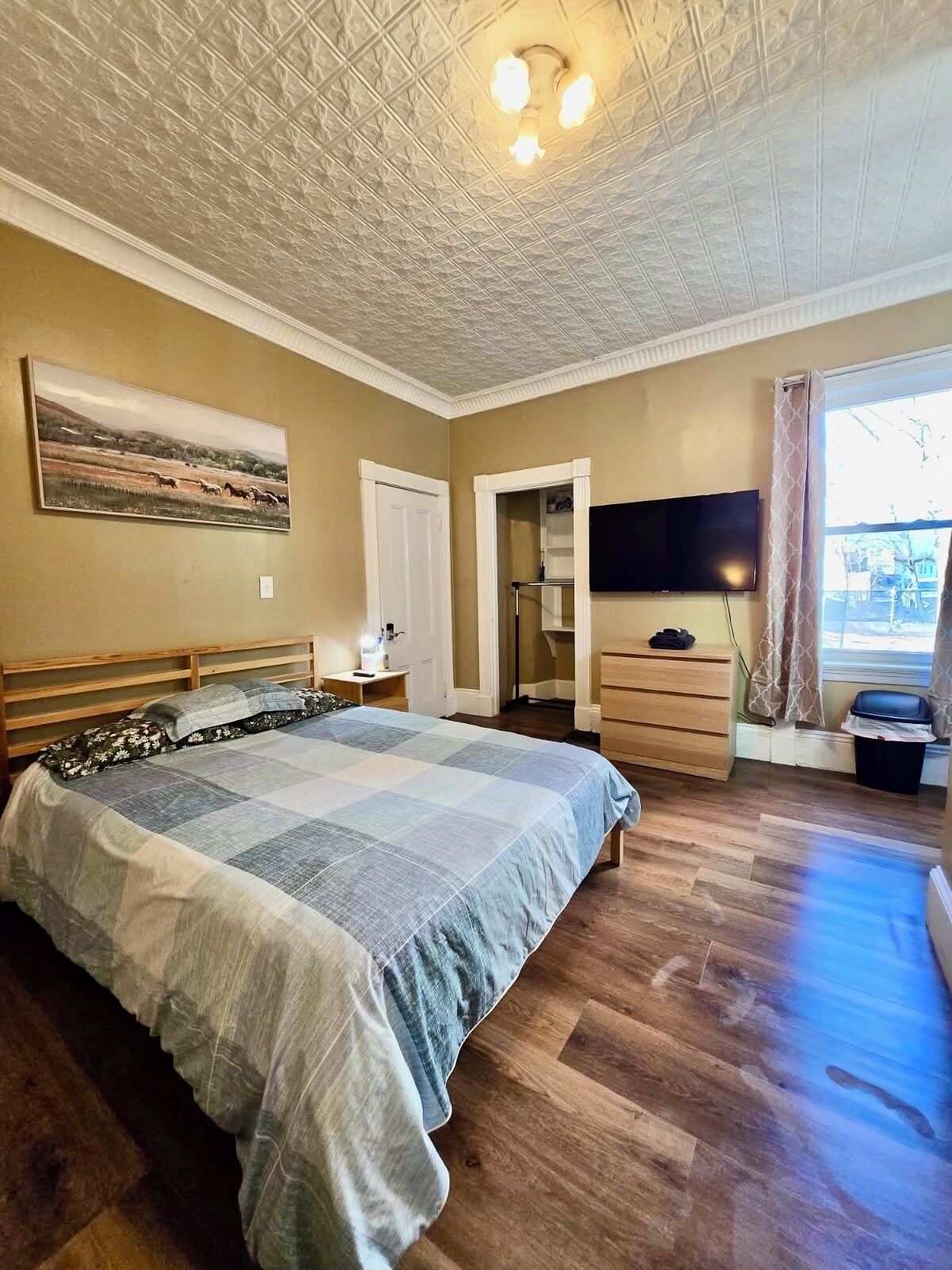 A bedroom with a bed, a television, a painting, and a window with curtains.