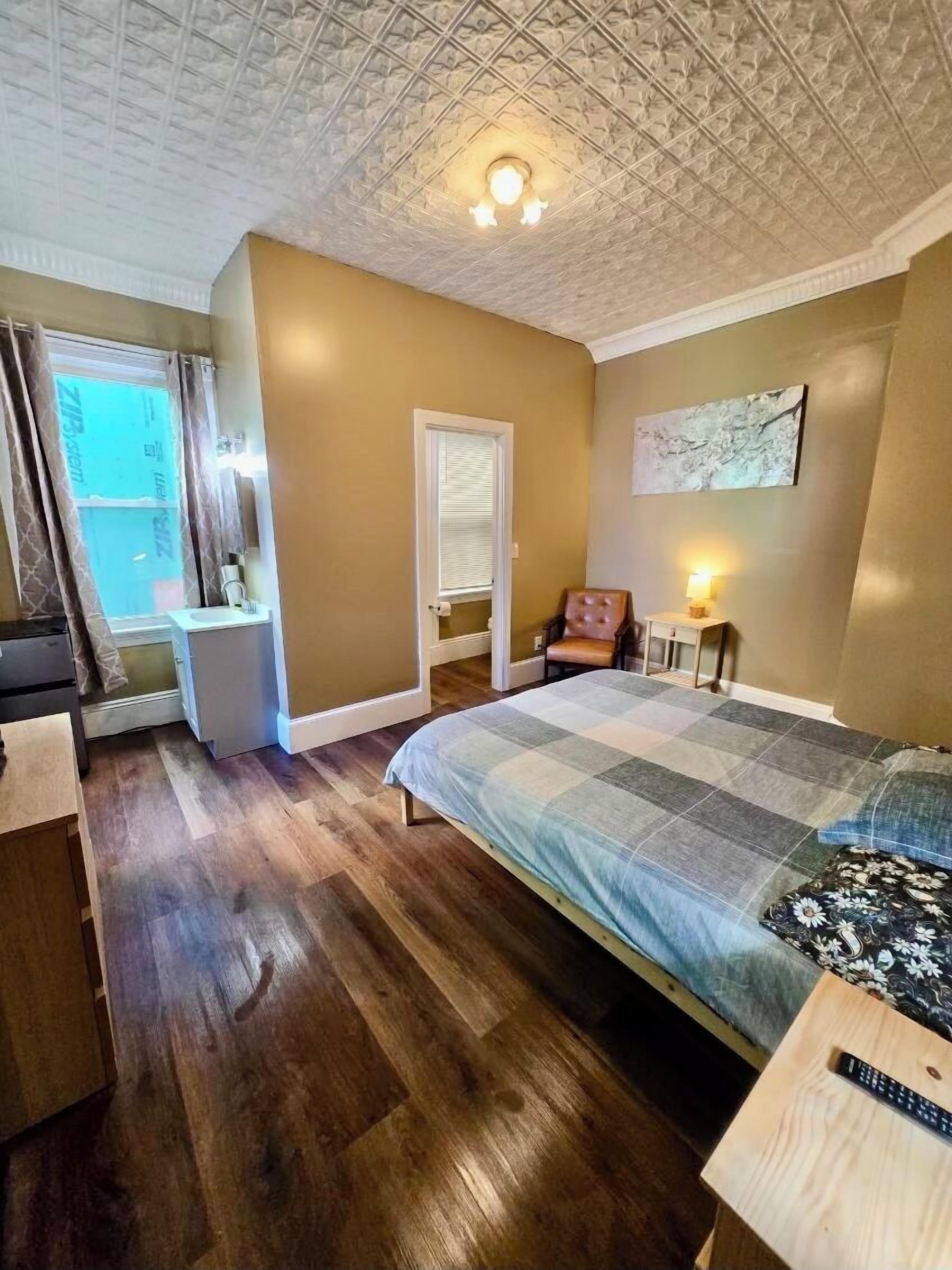 A bedroom with a bed, a chair, a nightstand, a window with curtains, and a ceiling light.