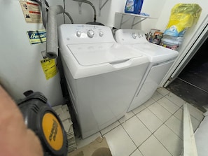 Eine Waschmaschine mit Frontlader und ein Trockner in einem Waschraum, mit sichtbaren Wasserrohren und einem gelben Warnhinweis.