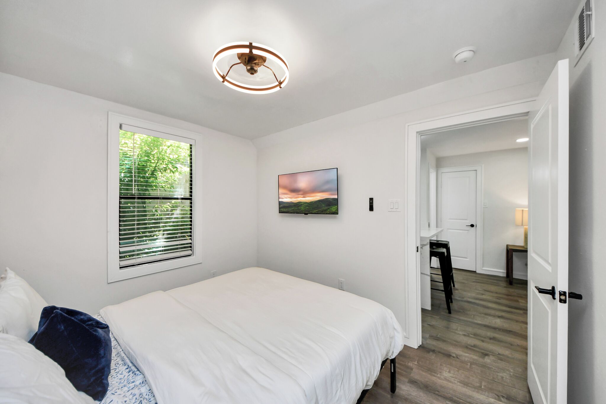 Une chambre à coucher avec un lit, une fenêtre avec des stores, un tableau accroché au mur et un couloir menant à une autre pièce.