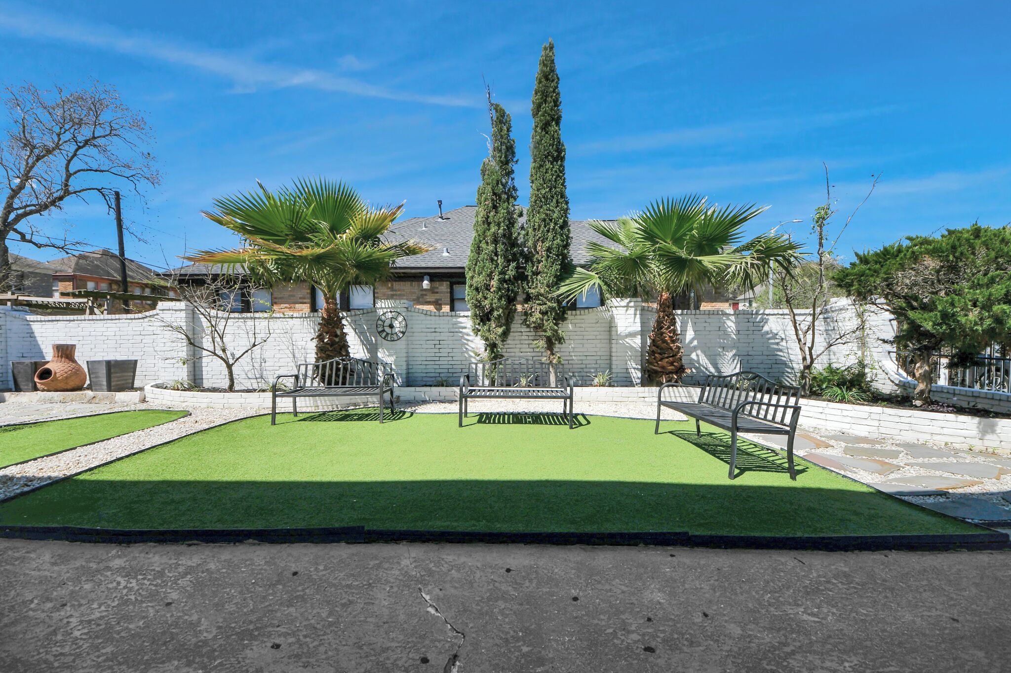 A courtyard with artificial grass, two benches, palm trees, and a white brick wall.