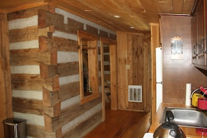 A log cabin interior with a wooden staircase, a kitchen area with a sink and kettle, and a window with a view.