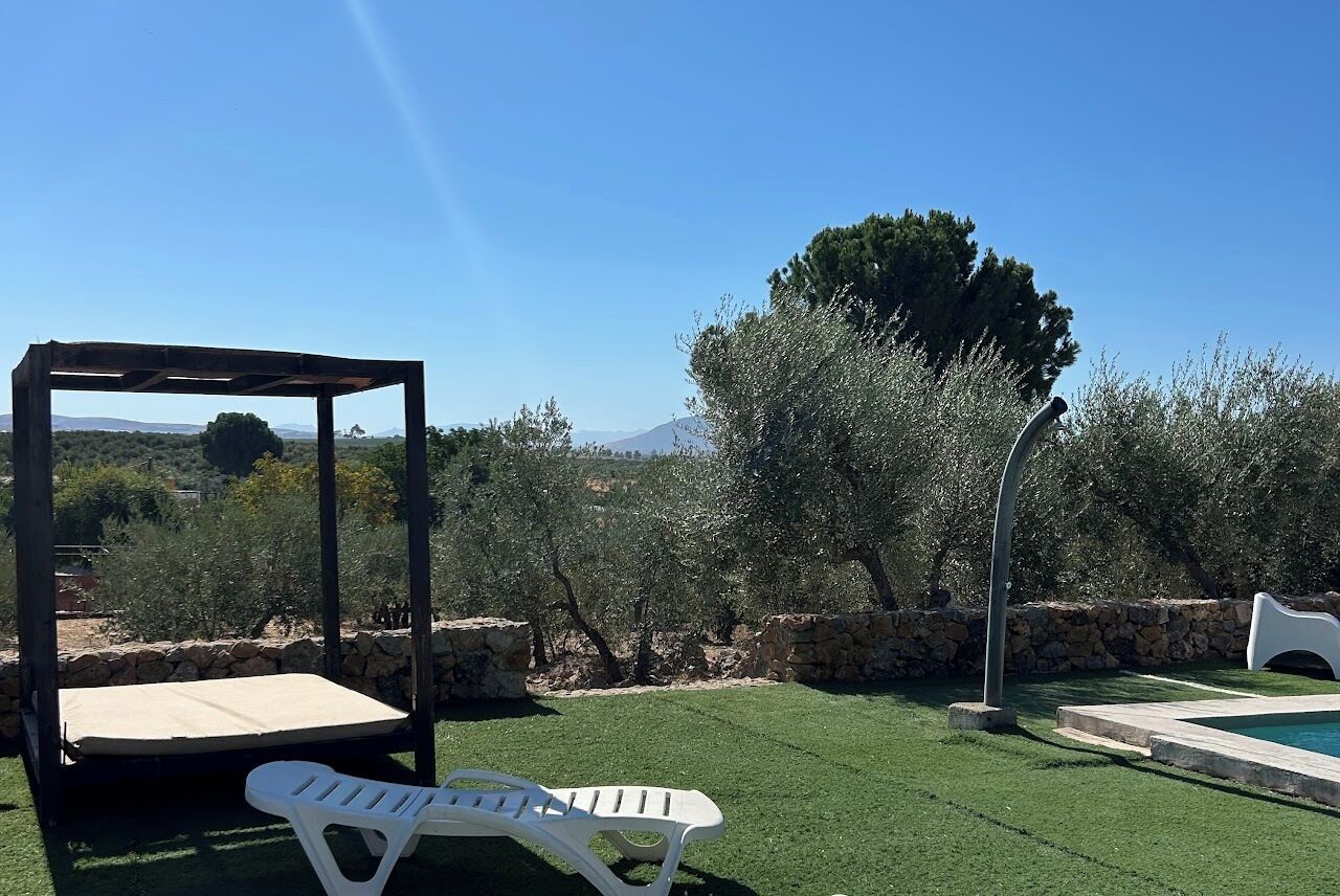 A gazebo with a mattress, a white lounge chair, a swimming pool, and a stone wall.