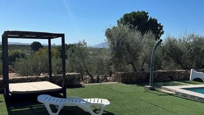 A gazebo with a mattress, a white lounge chair, a swimming pool, and a stone wall.