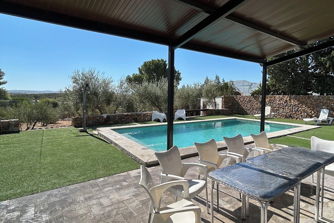 A covered outdoor area with a swimming pool, white plastic chairs, and a table.