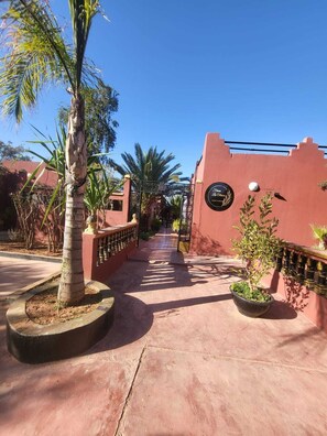 A pathway leading to a building with a red facade, palm trees, and a sign with a logo. - "Ferme des Oliviers" Guest House (Marrakesh)