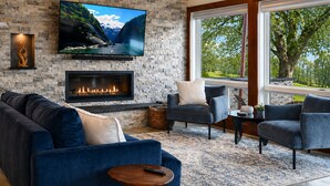 A living room with a fireplace, a TV mounted on a stone wall, and a large window with a view of greenery. - Serene | Hot Tub | Seneca Lake Access | Seneca Wine Trail (Geneva)