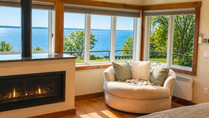 A cozy room with a fireplace, a cushioned seating area, and large windows offering a view of the sea. - Serene | Hot Tub | Seneca Lake Access | Seneca Wine Trail (Geneva)