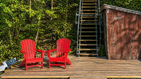 Une terrasse en bois avec deux fauteuils Adirondack rouges, une échelle en métal et un abri de jardin en bois.