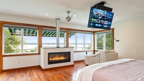 A modern bedroom with a fireplace, large windows, a ceiling fan, and a TV mounted on the wall. - Serene | Hot Tub | Seneca Lake Access | Seneca Wine Trail (Geneva)