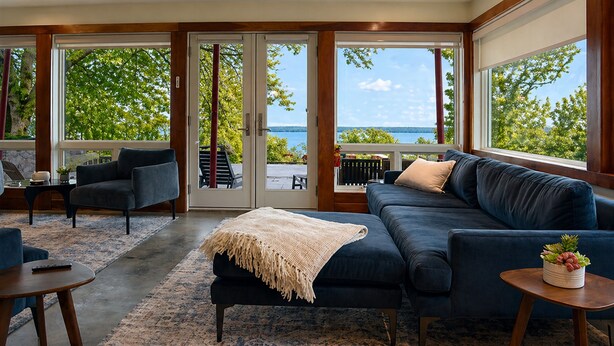 A modern living room with a dark blue sofa, a coffee table, and a view of the sea through large glass doors. - Serene | Hot Tub | Seneca Lake Access | Seneca Wine Trail (Geneva)