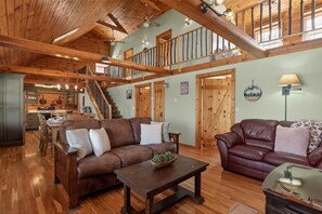 A spacious living area with a wooden ceiling, a staircase, and a kitchen in the background.