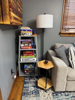 Uma sala de estar com um sofá cinza, almofadas estampadas, uma pequena mesa de madeira com uma luminária e uma estante cheia de jogos de tabuleiro.
