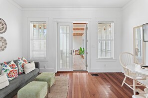 Una habitación con un sofá gris, otomanas verdes, una mesa de comedor blanca con una silla, y una puerta que da a un área exterior con plantas.