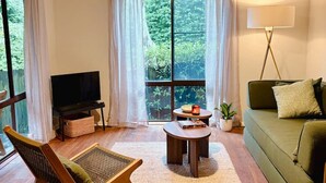 Uma sala de estar com sofá, poltrona, mesa de centro, televisão, abajur e uma janela grande com cortinas.