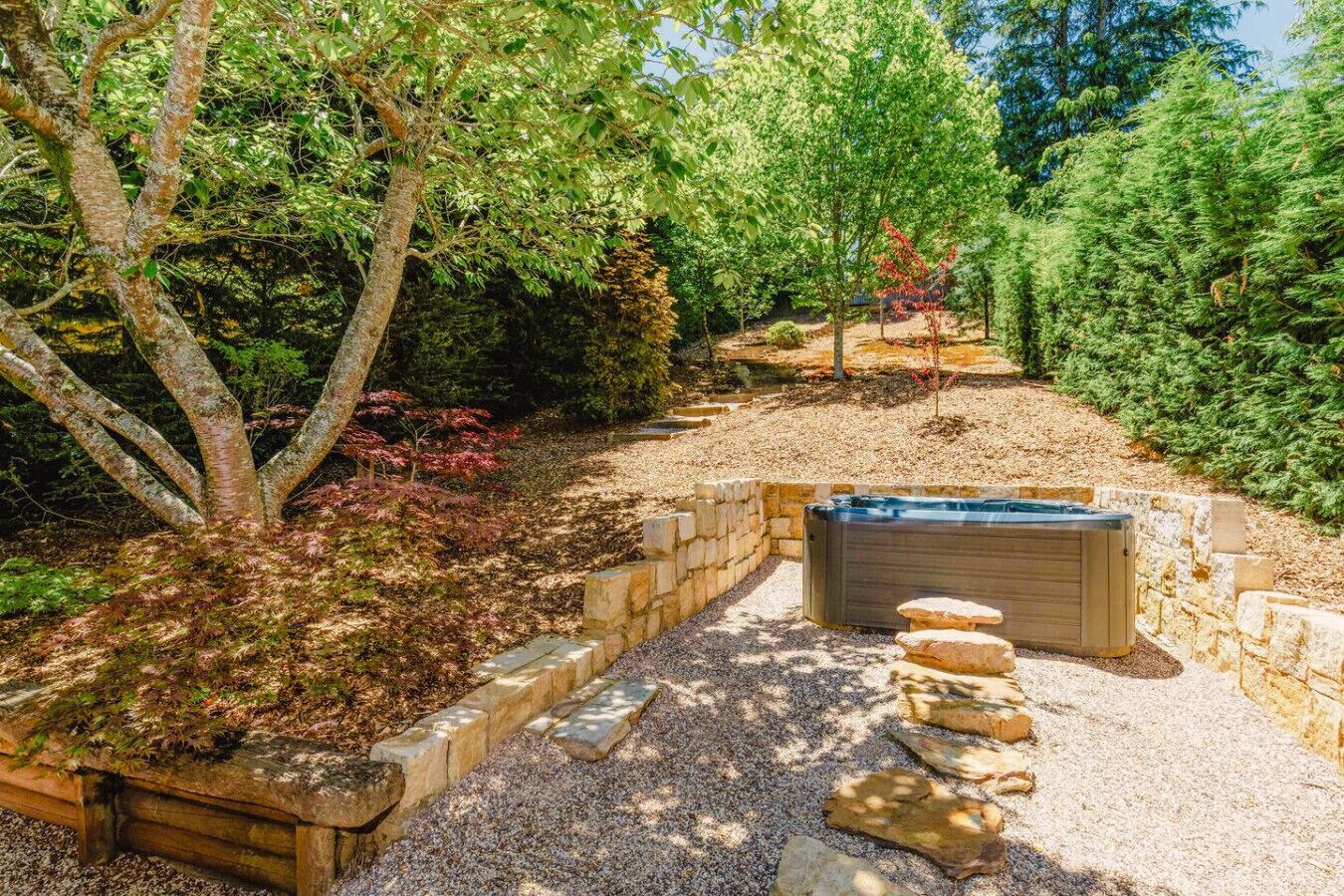 A hot tub surrounded by stone steps and a gravel path, with trees and shrubs in the background.