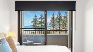 A hotel room with a bed, a balcony with a view of trees and the sea, and a table with chairs. - Happy Valley Tide - Bulcock Beachfront Apartment (Caloundra Region)