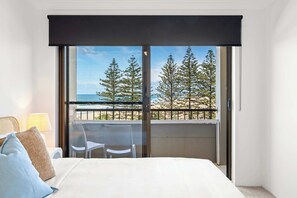 Une chambre d’hôtel avec un lit, un balcon donnant sur les arbres et la mer, et une table avec des chaises.