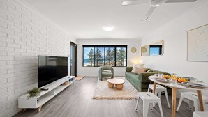 A modern living room with a flat-screen TV, a green sofa, a dining table with chairs, and a large window with a view of trees. - Happy Valley Tide - Bulcock Beachfront Apartment (Caloundra Region)