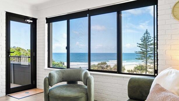 A room with a large window offering a view of the ocean and a white brick wall. - Happy Valley Tide - Bulcock Beachfront Apartment (Caloundra Region)