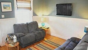 A living room with a grey sofa, a striped rug, a small side table with a lamp, and a television on the wall. - Renovated 3BR Shore House • Yard, Porch & Bay Breezes • Pet Friendly (Ocean City)