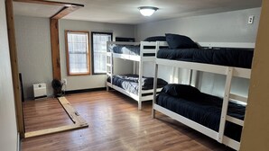 A room with wooden flooring, bunk beds, a ceiling fan, and a window with blinds.