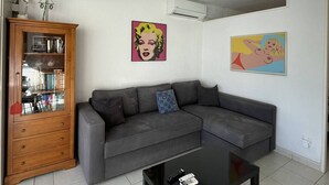 Ein Wohnzimmer mit einem grauen Ecksofa, einem Holzschrank, einem schwarzen Couchtisch und zwei Pop-Art-Gemälden an der Wand.