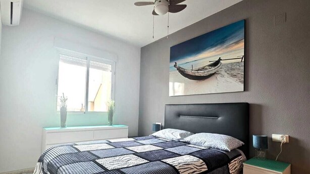 A bedroom with a bed, a ceiling fan, a window, a nightstand, a bedside table, and a painting. - Penthouse city sea and sun (Torrevieja)