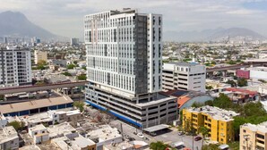 Un edificio de varios pisos con un diseño arquitectónico moderno, rodeado por un paisaje urbano con diferentes edificios y una montaña al fondo.