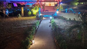Un chemin menant à un bâtiment doté d’un toit rouge, entouré de guirlandes lumineuses et de plantes décoratives.