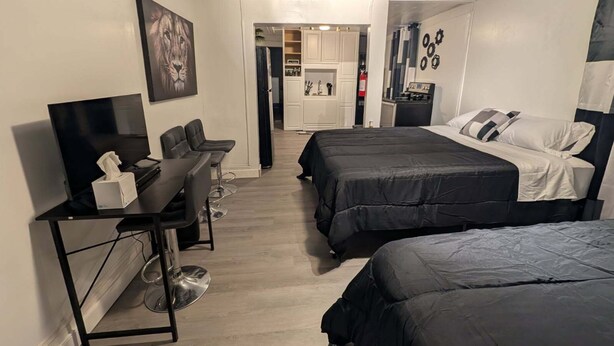 A hotel room with a TV, two beds, a desk with a chair, and a kitchenette in the background.
