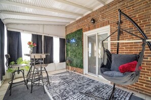 Une pièce avec un fauteuil suspendu, une table entourée de chaises, un tapis à motifs et un mur de briques orné de plantes grimpantes.