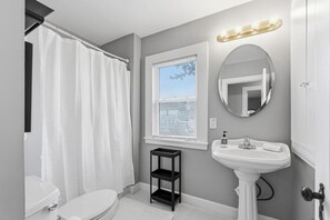 Une salle de bain comprenant des toilettes, un lavabo, un miroir, un rideau de douche et une fenêtre avec des stores.