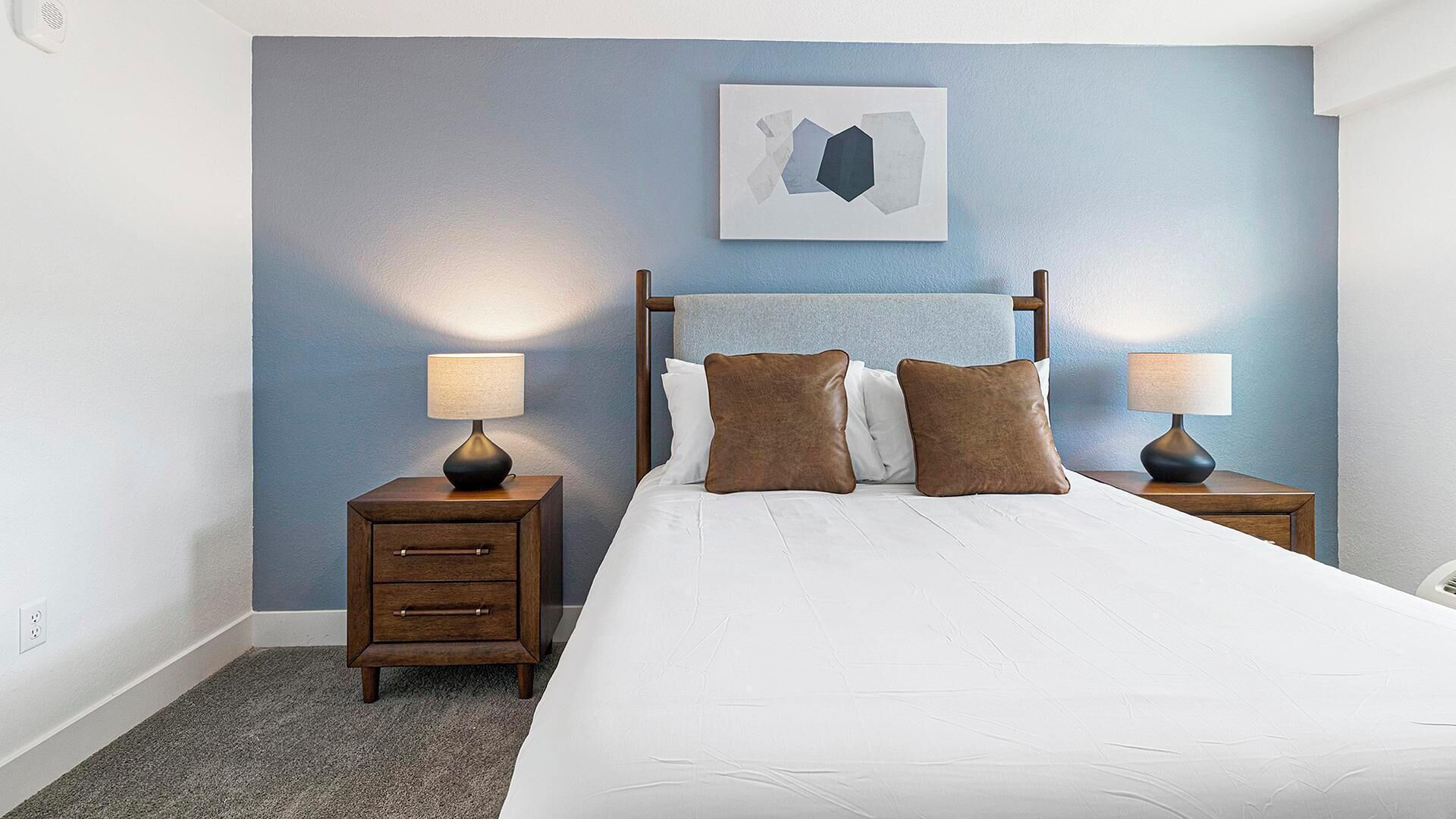 Una habitación de hotel con una cama, dos mesitas de noche, una mesita auxiliar, un cuadro en la pared y suelo alfombrado.
