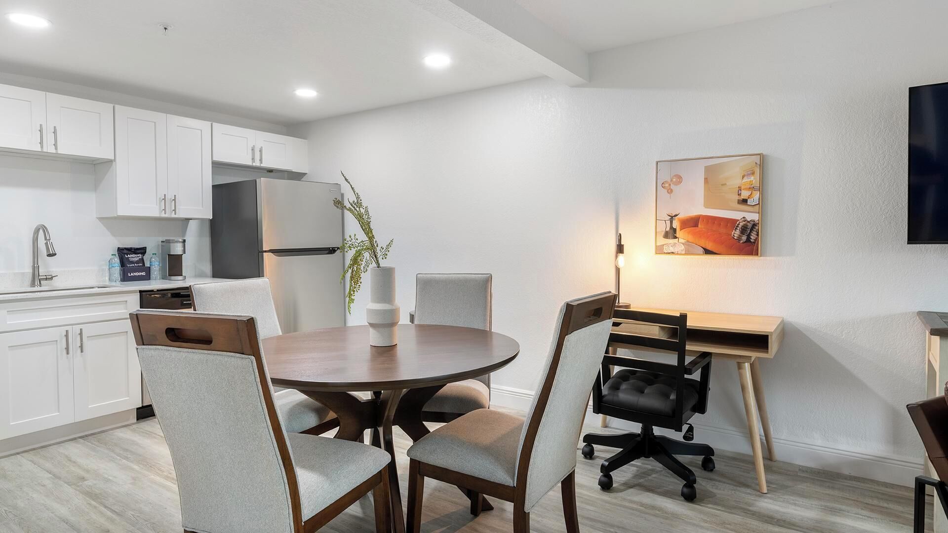 A modern kitchen with white cabinets, a refrigerator, and a coffee maker. There is a dining area with a round table and chairs, and a TV mounted on the wall.