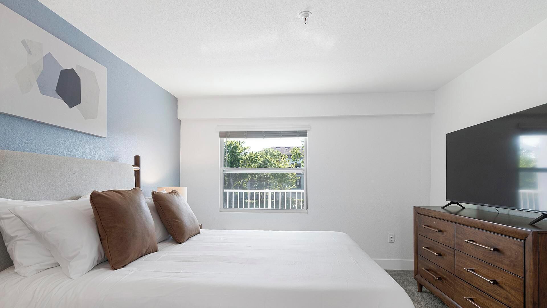 Ein Schlafzimmer mit Bett, Kommode, Fernseher, Fenster und Deckenleuchte.