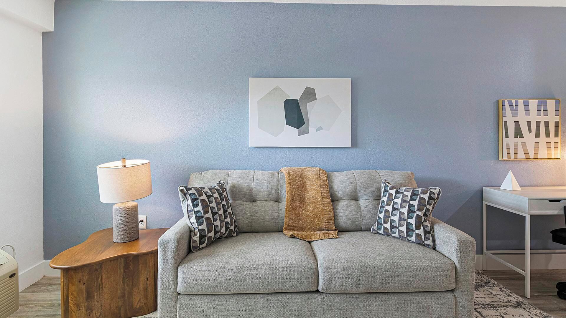 Sala de estar com sofá cinza, almofadas estampadas, manta, mesa lateral com abajur e arte abstrata na parede.