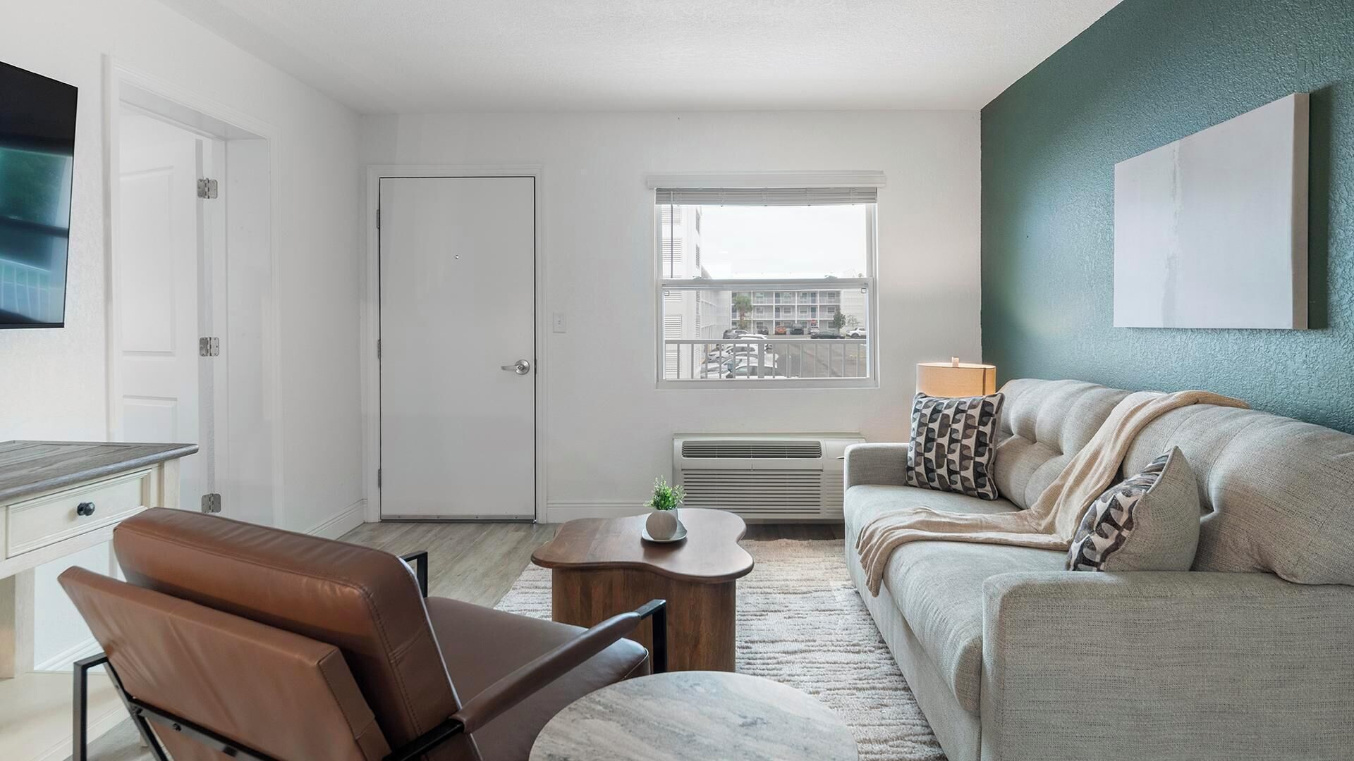 A modern living room with a sofa, a coffee table, a brown leather chair, a white door, and a window with a city view.