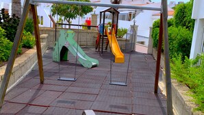 Un parque infantil con columpios y toboganes, rodeado de vegetación y edificios.