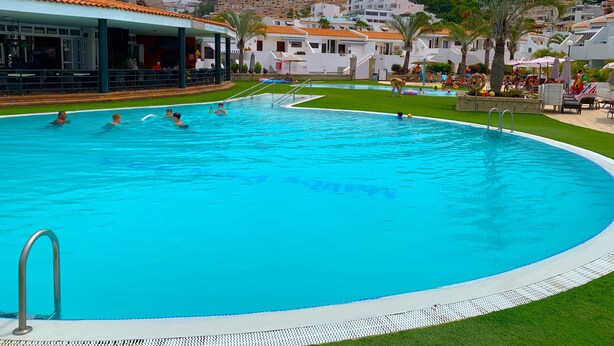 Una piscina con un deck perimetral, un edificio con techo de tejas y una colina con edificaciones residenciales al fondo.