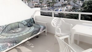 Un balcón con una cama, sillas blancas y vistas a una población densamente poblada con palmeras.