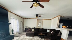 A living room with a sectional sofa, a ceiling fan, a fireplace, and a kitchen area in the background. - Quiet Country Getaway ,quiet, and spacious getaway (Belen)