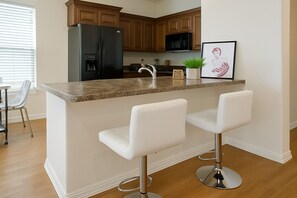 Una cocina con gabinetes de madera, un refrigerador negro, un microondas, un lavamanos y dos taburetes blancos de bar.