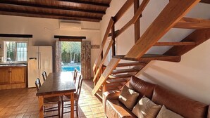 A rustic interior with a wooden staircase, a dining area with a table and chairs, a kitchen with a refrigerator, and a view of a swimming pool.