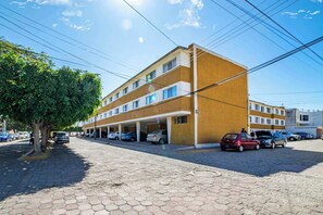 A multi-story building with a parking garage, a tree-lined street, and parked cars. - +MS +Única Suite +Inmejorable ubicación +Internet (Santiago de Querétaro)