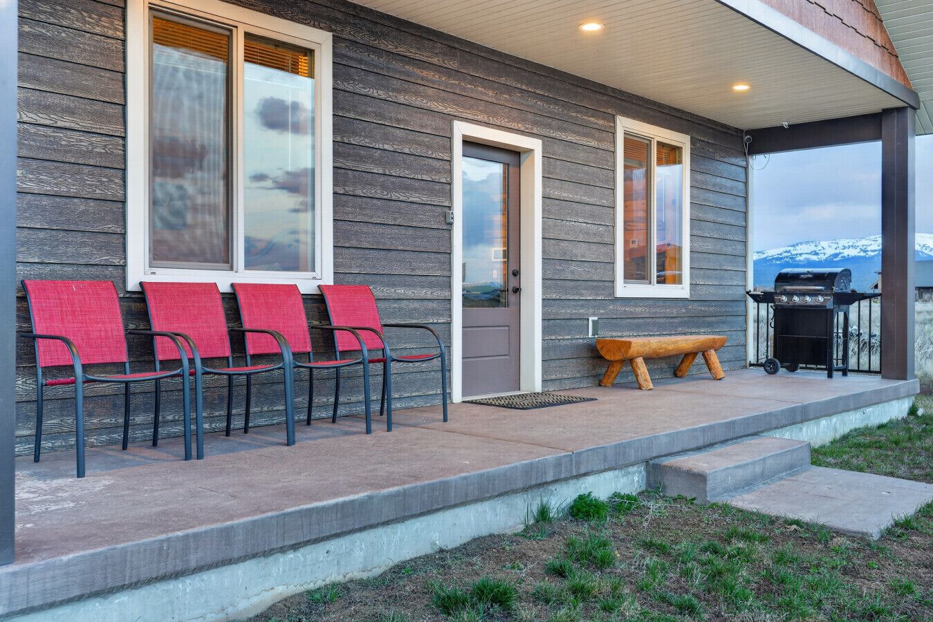 A building with a porch, four red chairs, a bench, and a barbecue grill.